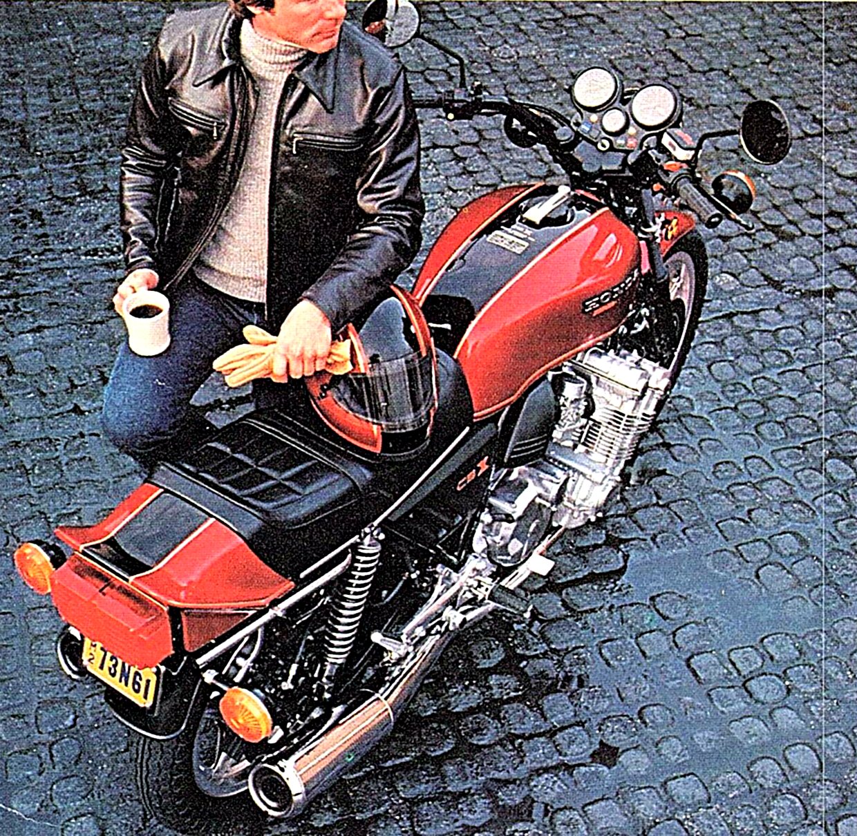 A rider in a black leather jacket sits on a CBX holding a cup of coffee on wet cobblestones.