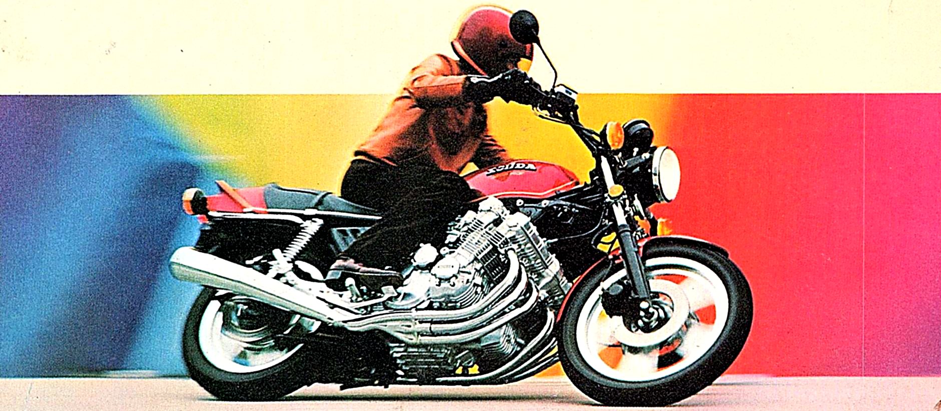 A rider leans a red Honda CBX through a turn in front of a rainbow pop-art backdrop.