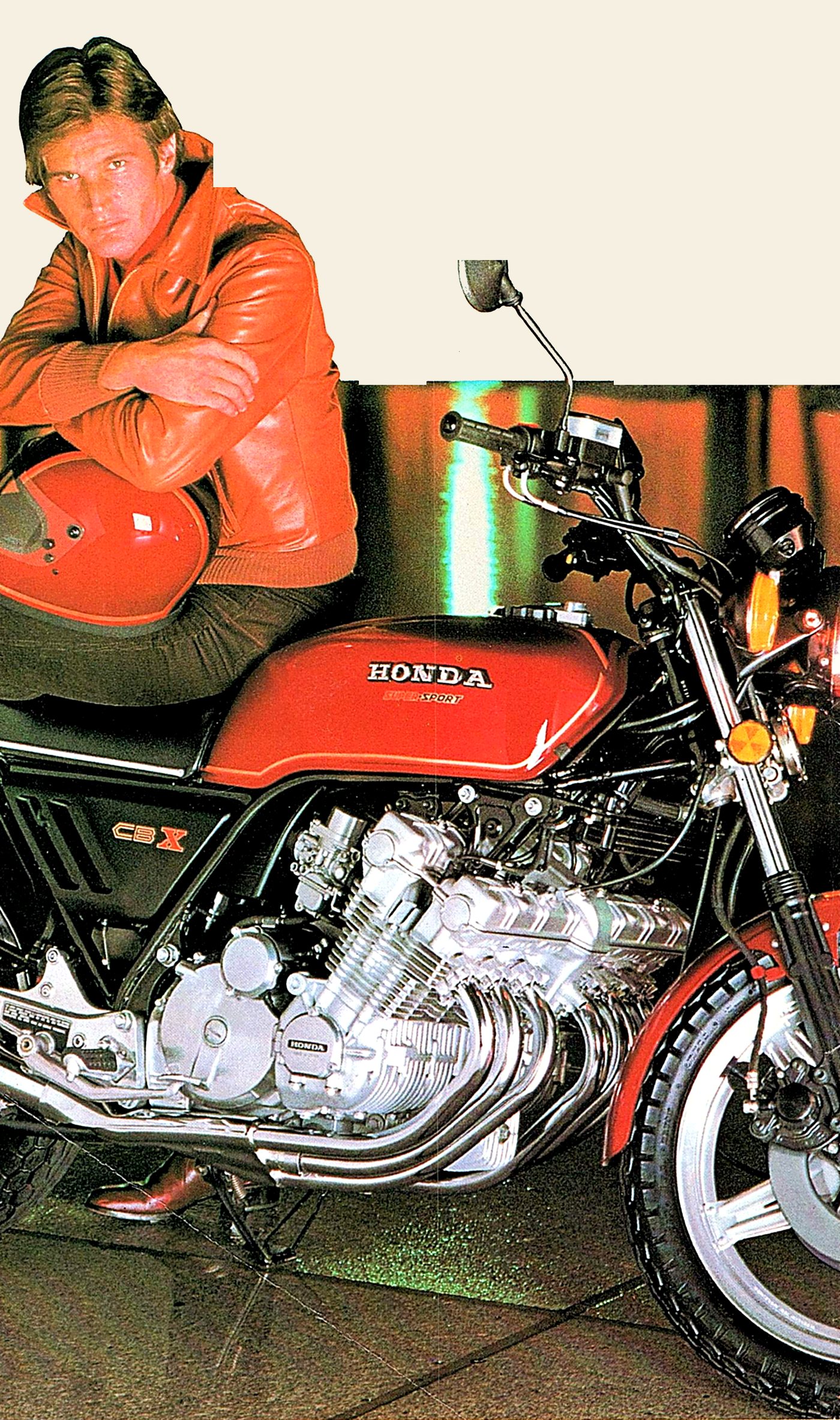 A man in an orange leather jacket leans on a red 1979 Honda CBX, its six-cylinder engine gleaming in a dark studio.